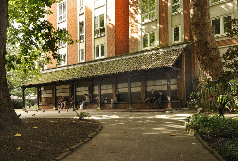 Visitors seated beneath the timber shelter of the Watts Memorial, with rows of ceramic tablets behind them and a shaded path in front.