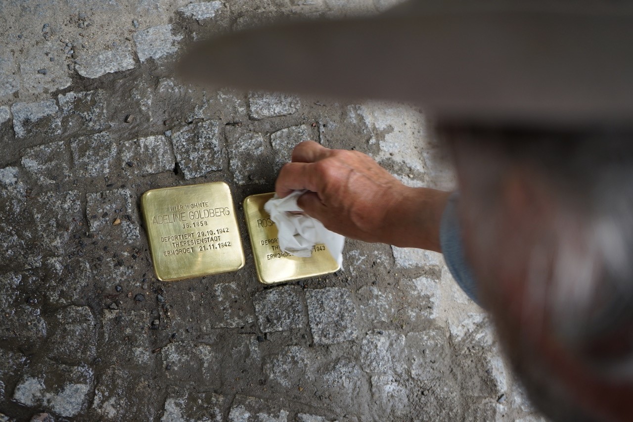 An overhead photograph of Gunter Demnig wiping a brass Stolperstein beside the memorial for Adeline Goldberg, with both stones set into the pavement.