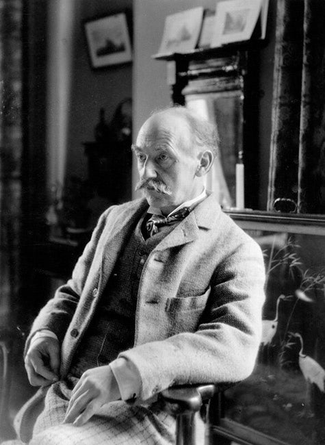 Archival photograph of Thomas Hardy seated in a light jacket and dark waistcoat, with books and framed pictures behind him.