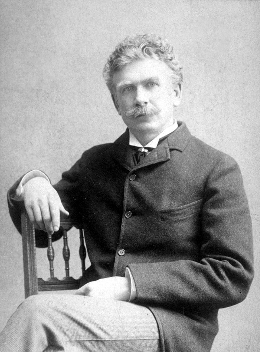 Archival photograph of Ambrose Bierce seated sideways on a wooden chair, with a moustache, dark jacket, and light trousers.