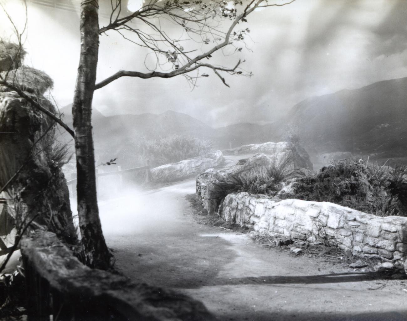 Black-and-white archival set photograph of a winding Highland road built in a film studio, with painted mountain scenery visible beyond it.