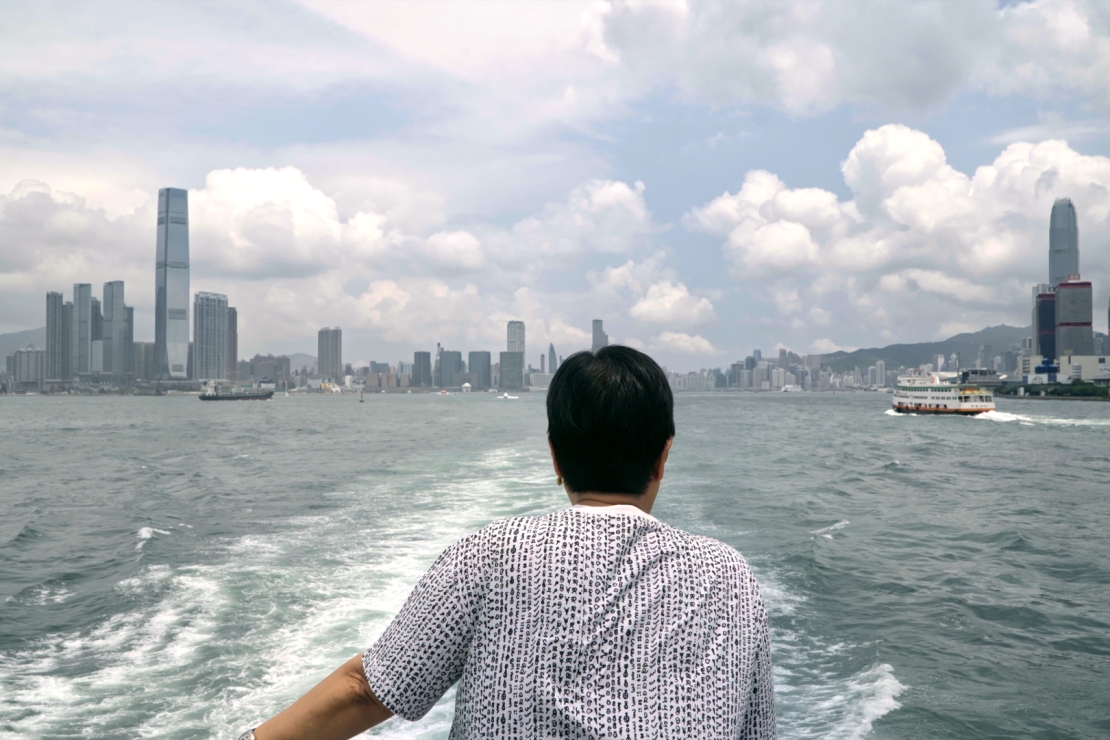 Ann Hui seen from behind aboard a ferry, looking across Victoria Harbour at Hong Kong's skyline, in a publicity still from Keep Rolling.