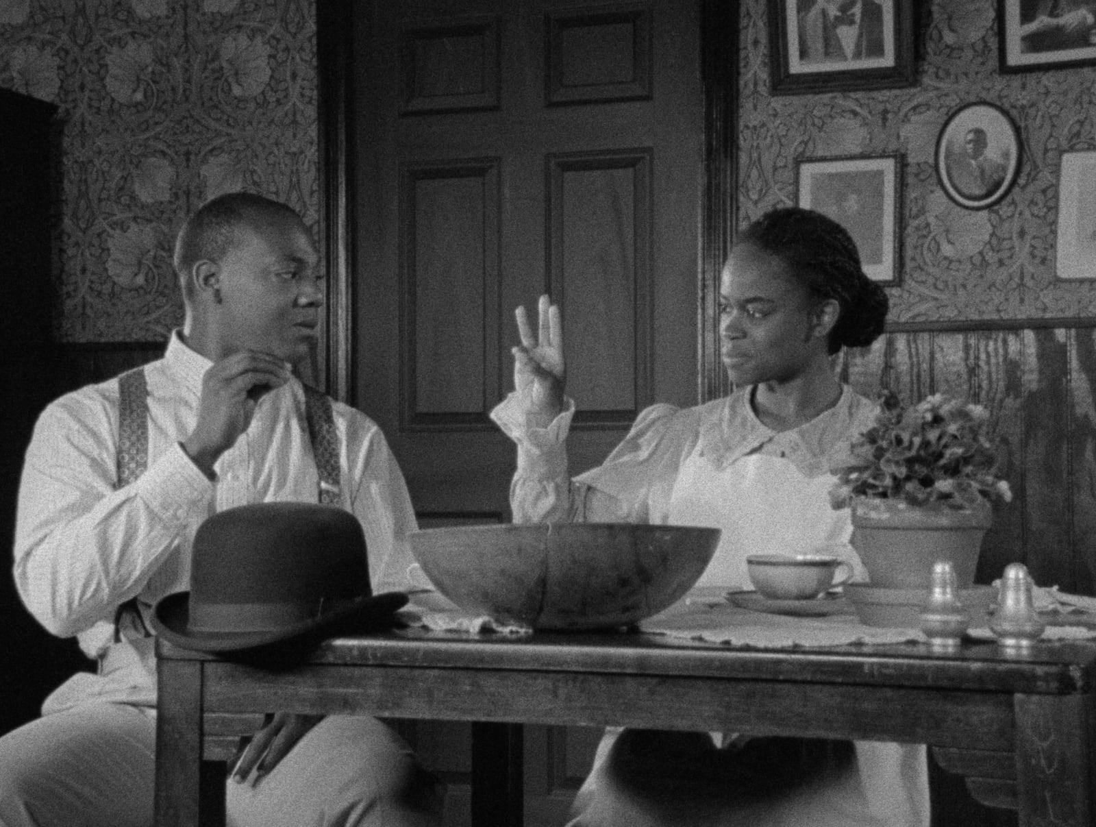 Black-and-white film still of John Earl Jelks and Michelle A. Banks seated at a dining table, watching each other's hands as they sign.