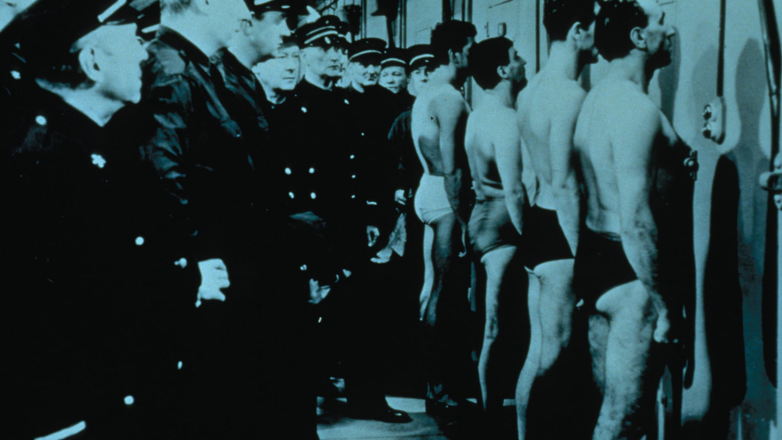 Prisoners in underwear face cell doors while a group of uniformed guards fills the corridor in an archival still from Le Trou.