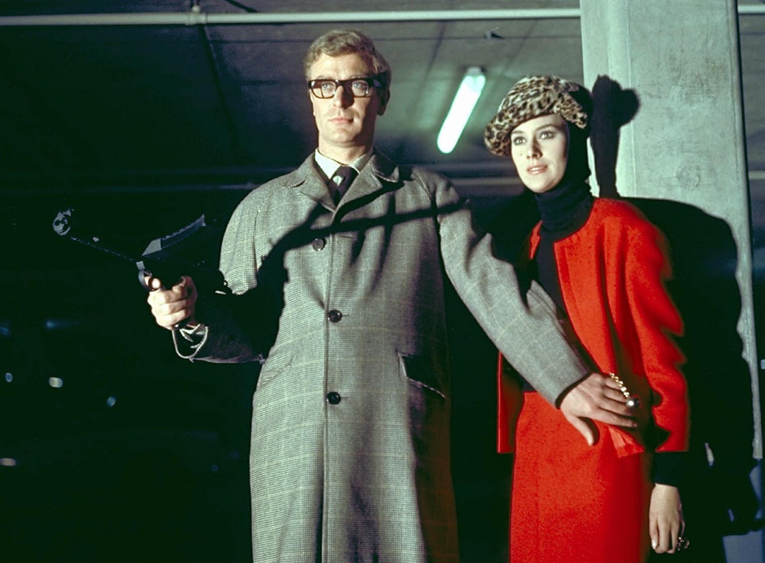 Michael Caine holds a submachine gun and shields Sue Lloyd, wearing a red coat, beside a concrete column in The Ipcress File.