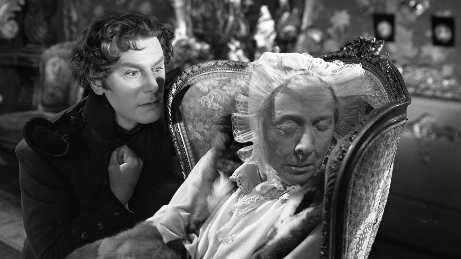 Black-and-white still from The Queen of Spades showing Herman Suvorin leaning beside Countess Ranevskaya in an ornate upholstered chair, with ornaments and reflective surfaces crowding the room behind them.