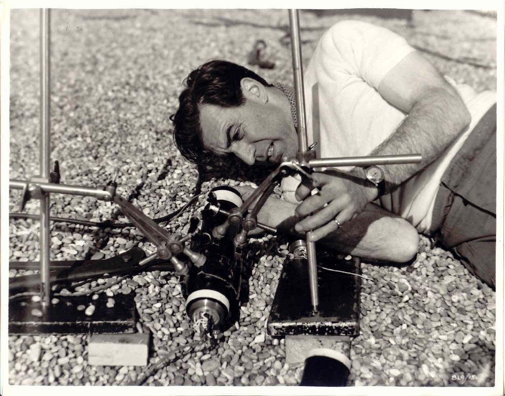 David Farrar as Sammy Rice lying on the shingle of Chesil Beach and working on a bomb held in metal stands, in a black-and-white studio still.