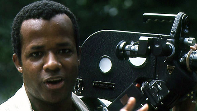 William Greaves holds a motion-picture camera beside his face, with trees behind him.