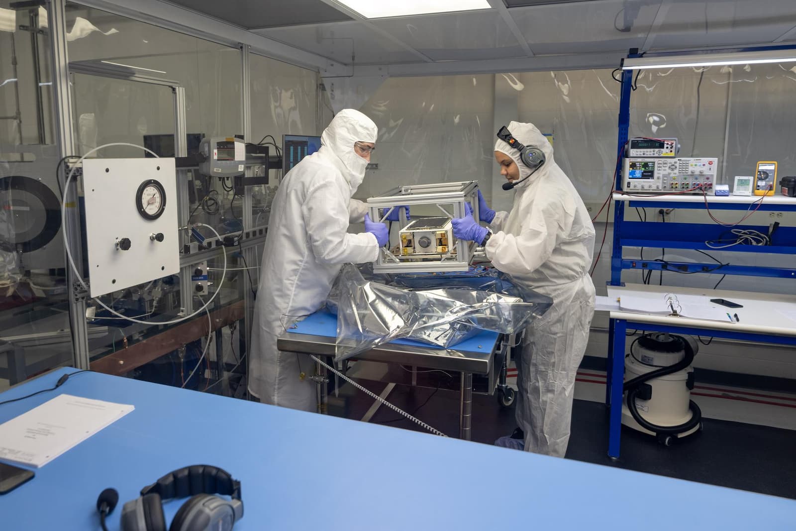 Two engineers in protective cleanroom clothing prepare a small spacecraft on a laboratory workbench.