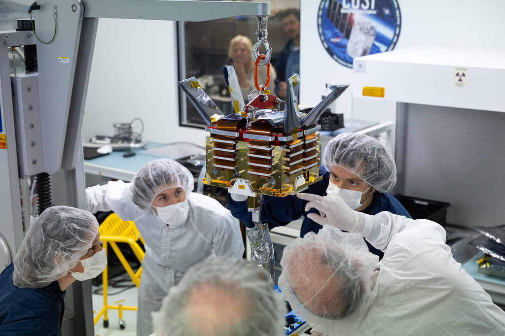 Engineers and managers in cleanroom clothing watch a gold and silver COSI detector assembly suspended above a laboratory table.