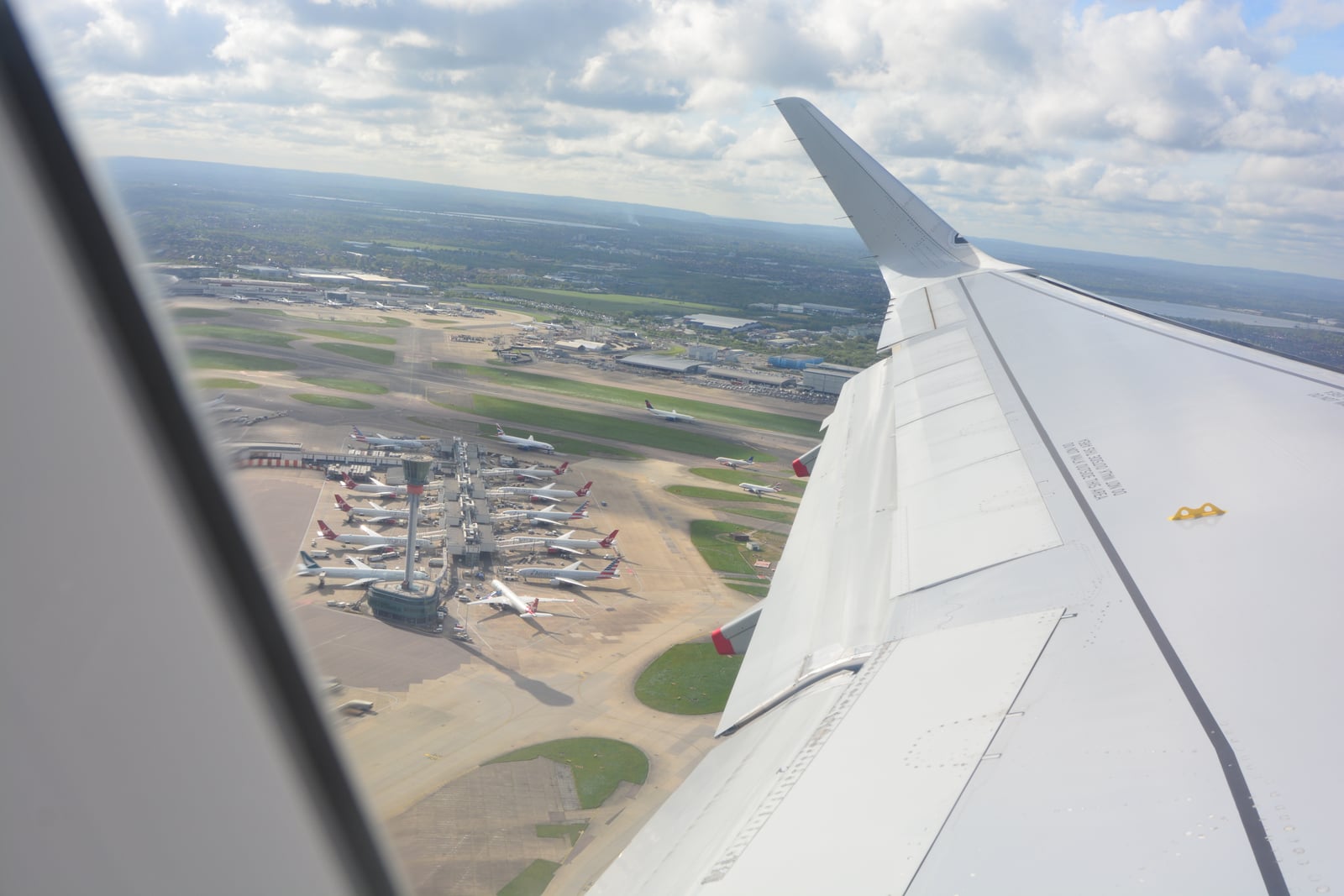 Heathrow's parked aircraft, taxiways and control tower seen through a passenger window, beside the wing of a departing aircraft.