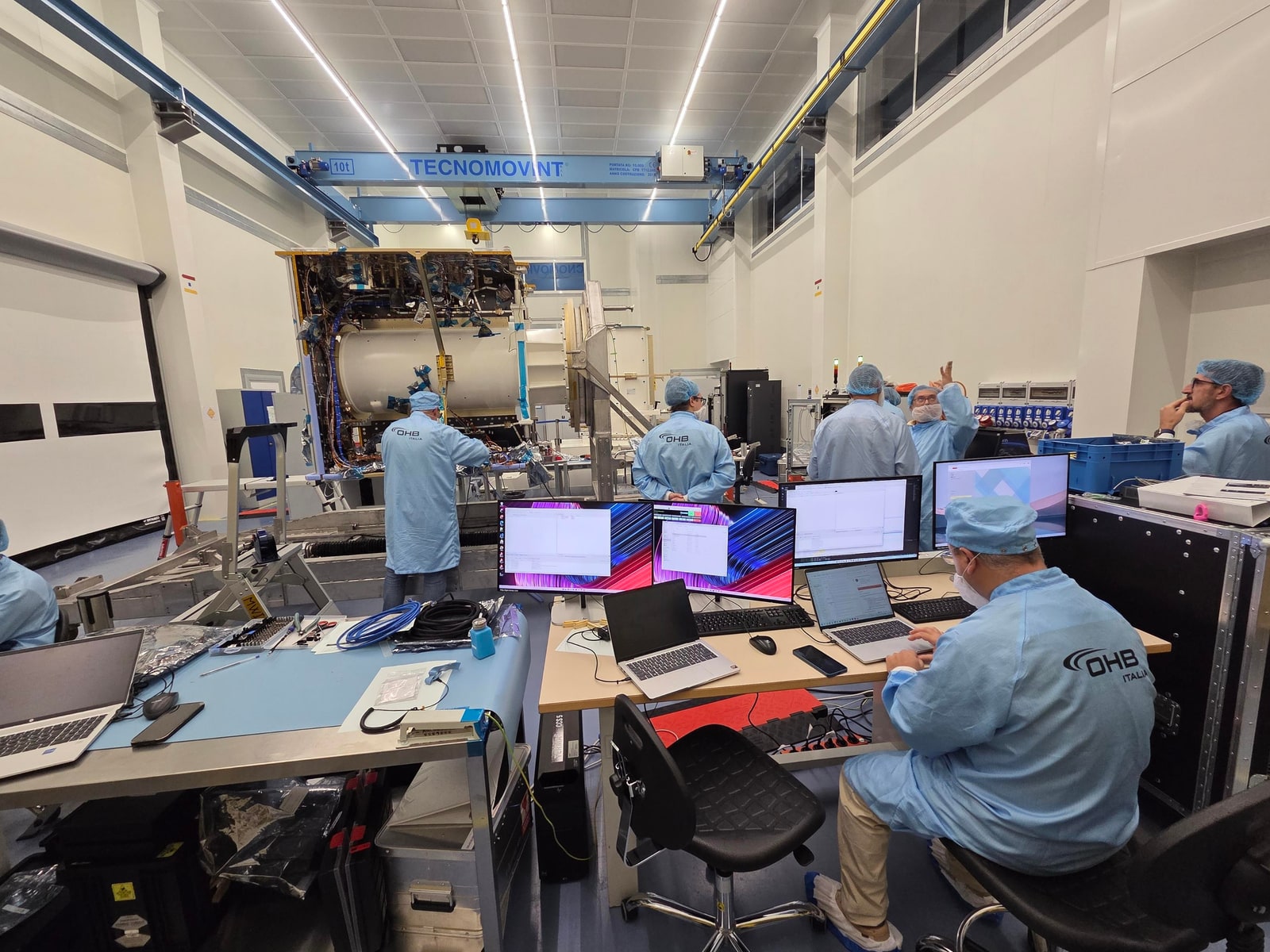 Engineers in blue cleanroom clothing monitor computers beside the open Ramses spacecraft at OHB Italia in Milan.