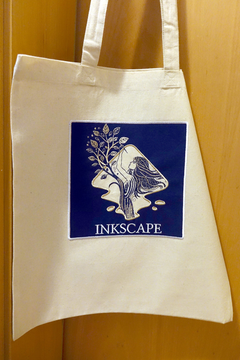 A cream tote bag hanging against a wooden door, with a blue embroidered patch showing the Inkscape design and a figure reaching toward leafy branches.