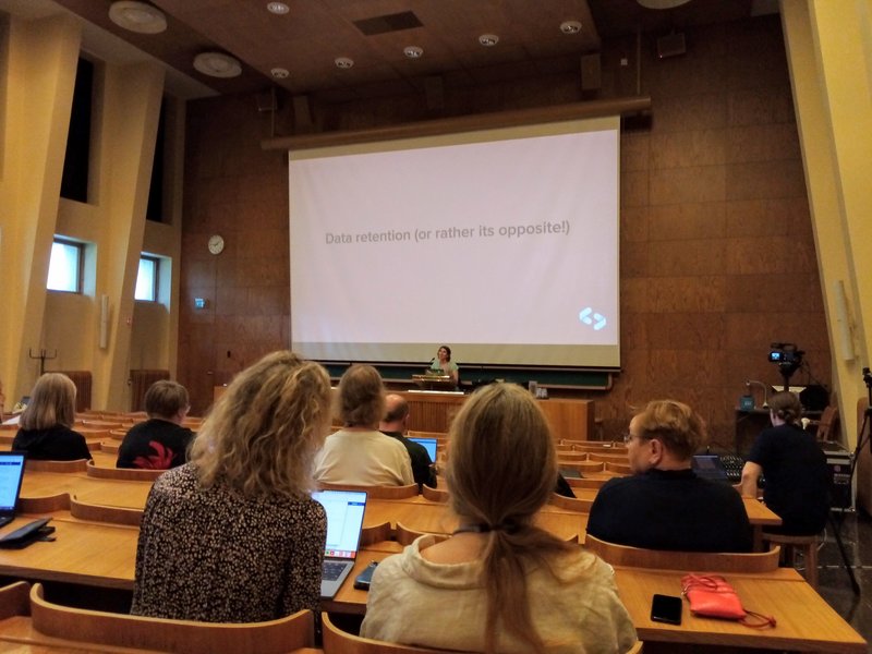 A speaker addresses KohaCon attendees in a wood-lined lecture hall while a presentation about patron data retention is projected.