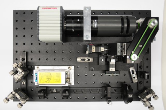 An OpenSPIM microscope assembled on a perforated optical breadboard, with a camera, lens assemblies, mirrors, and a central specimen stage.