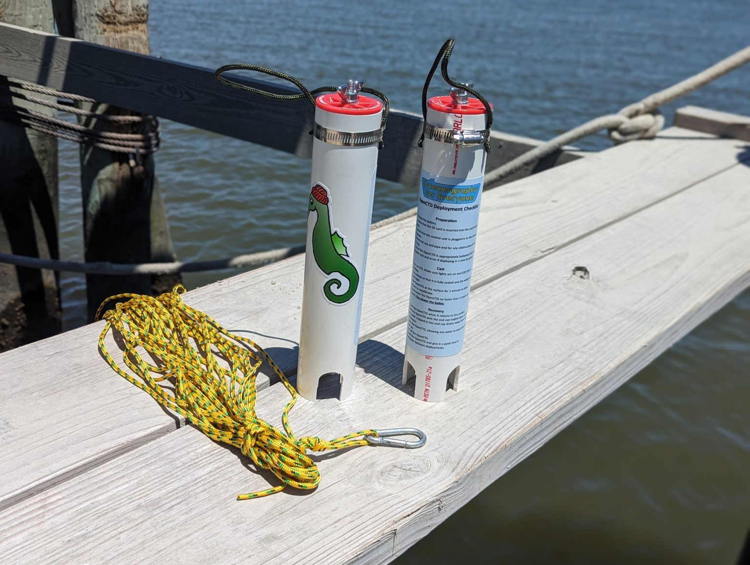 Two white OpenCTD instruments with red caps stand on a wooden dock beside a coiled yellow casting line and the water.