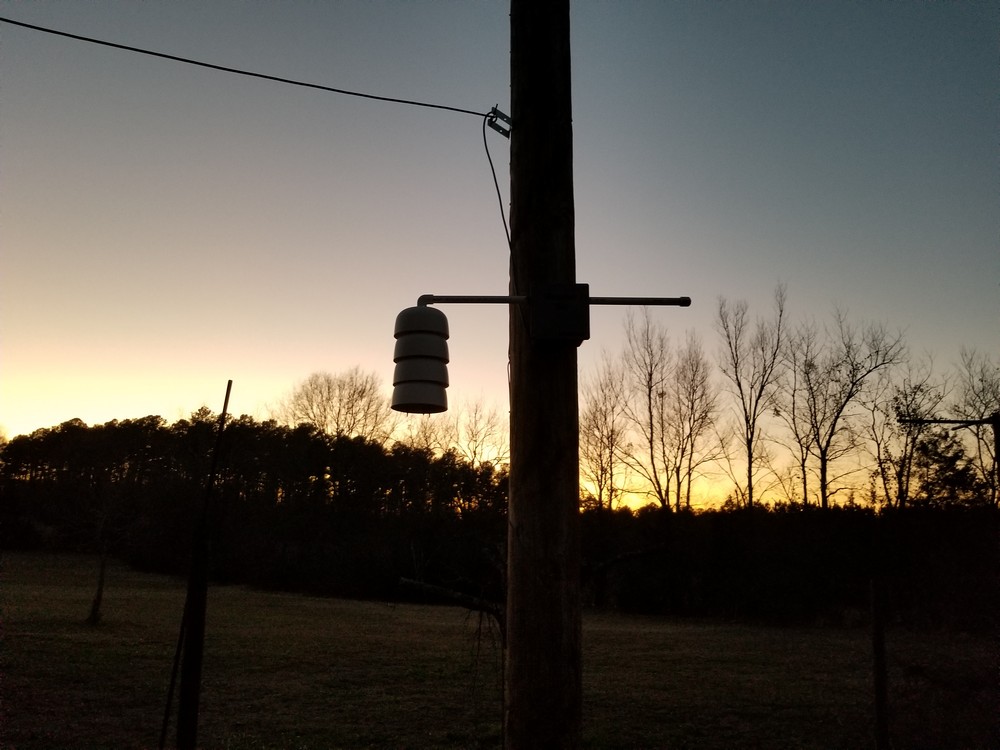 A weather sensor shield hangs from a horizontal pipe on a wooden support at dusk, above grass and a line of trees.