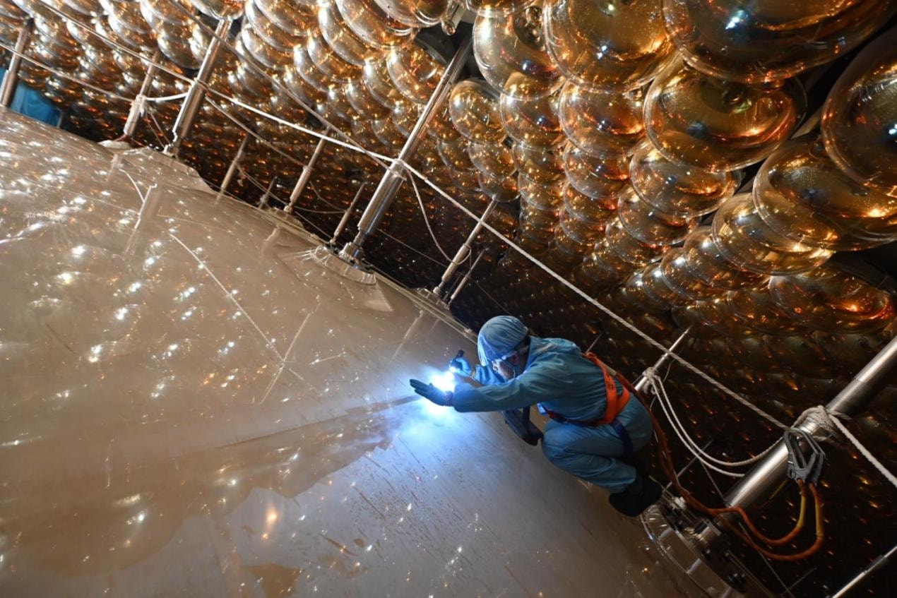 A technician in protective clothing examines JUNO's acrylic sphere beneath rows of gold-coloured photomultiplier tubes.