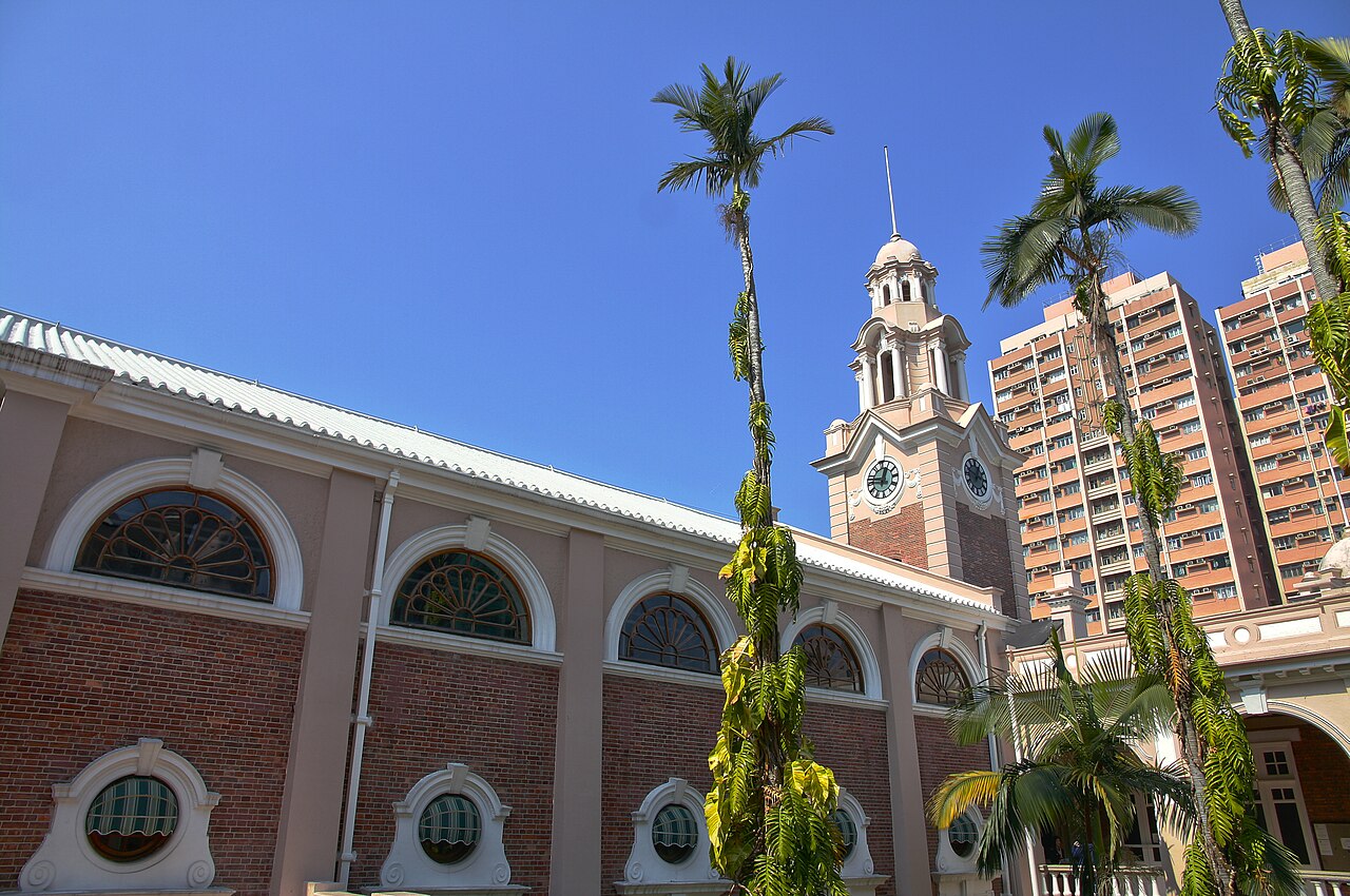A red-brick courtyard wall and clock tower at the University of Hong Kong, framed by tall palm trees under a blue sky.