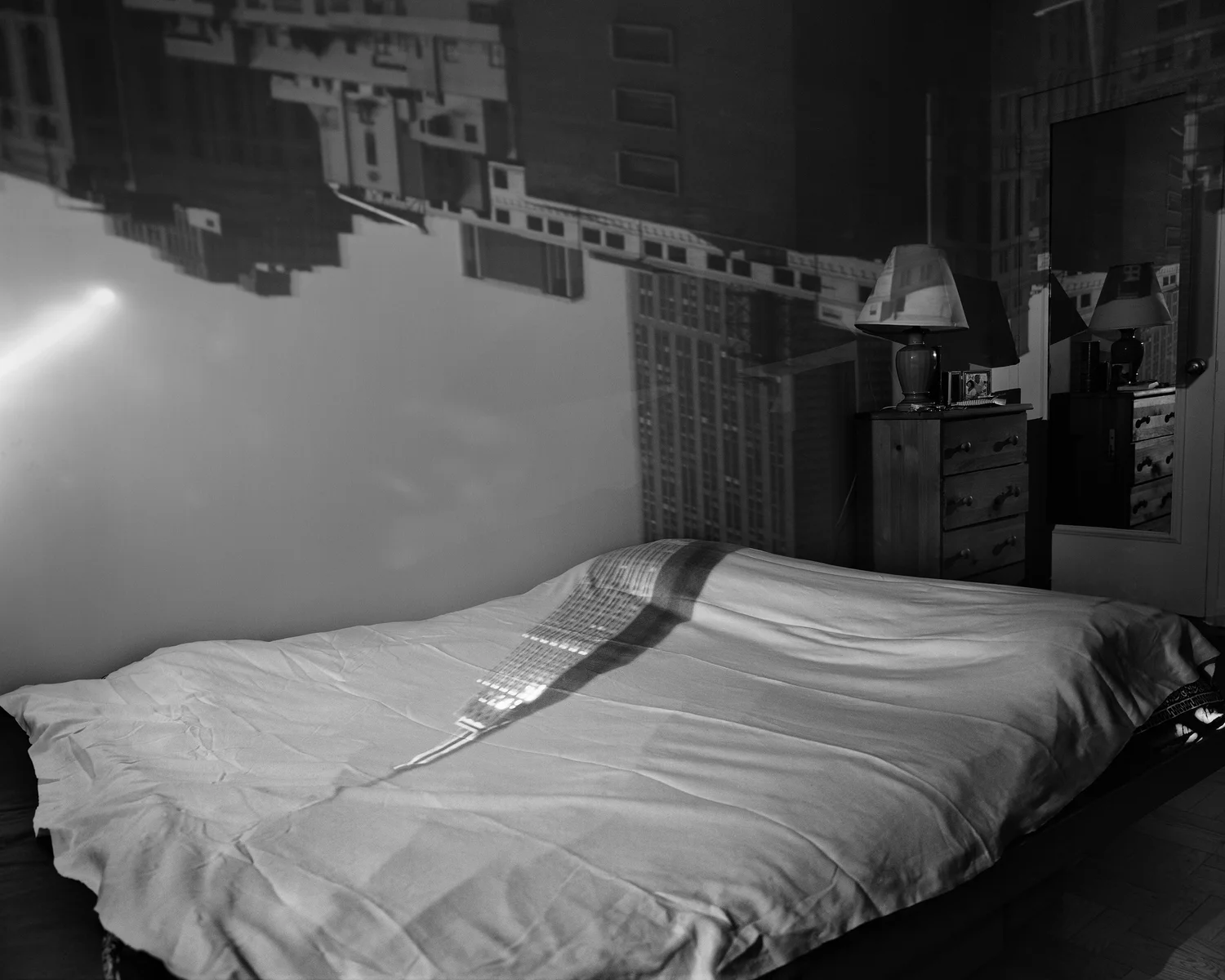 Black-and-white photograph of an inverted Empire State Building projected down a bedroom wall and across a bed, with a lamp, framed photograph, and mirror at right.