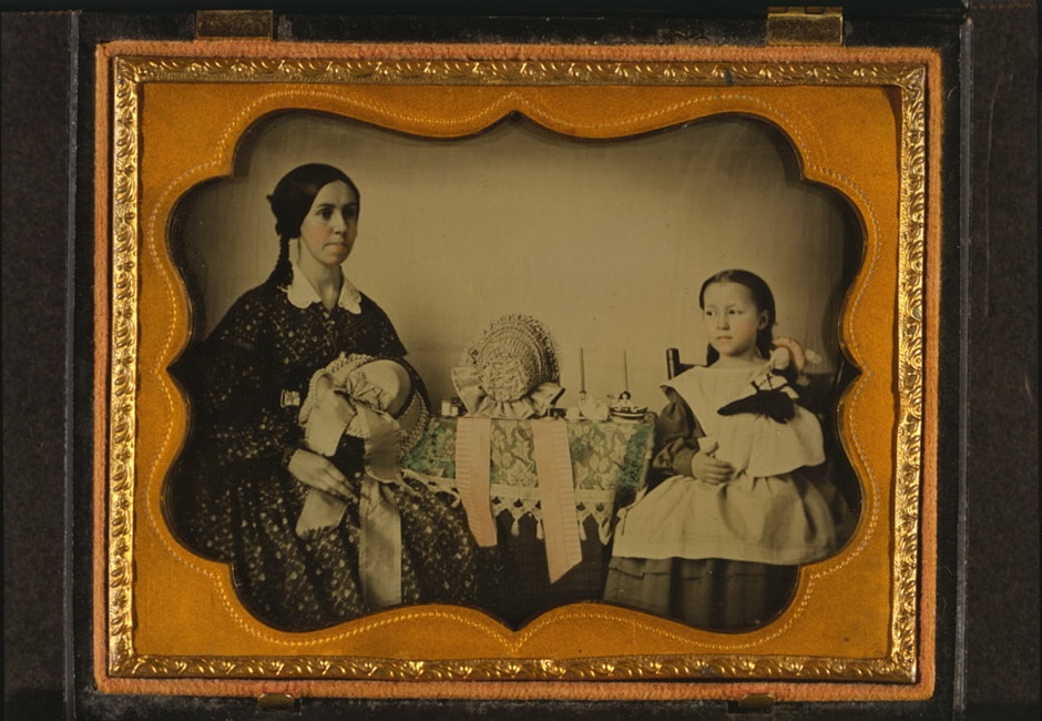 An early photograph of a woman and girl seated beside a table with bonnets and a doll, enclosed by an ornate gold-colored mat.