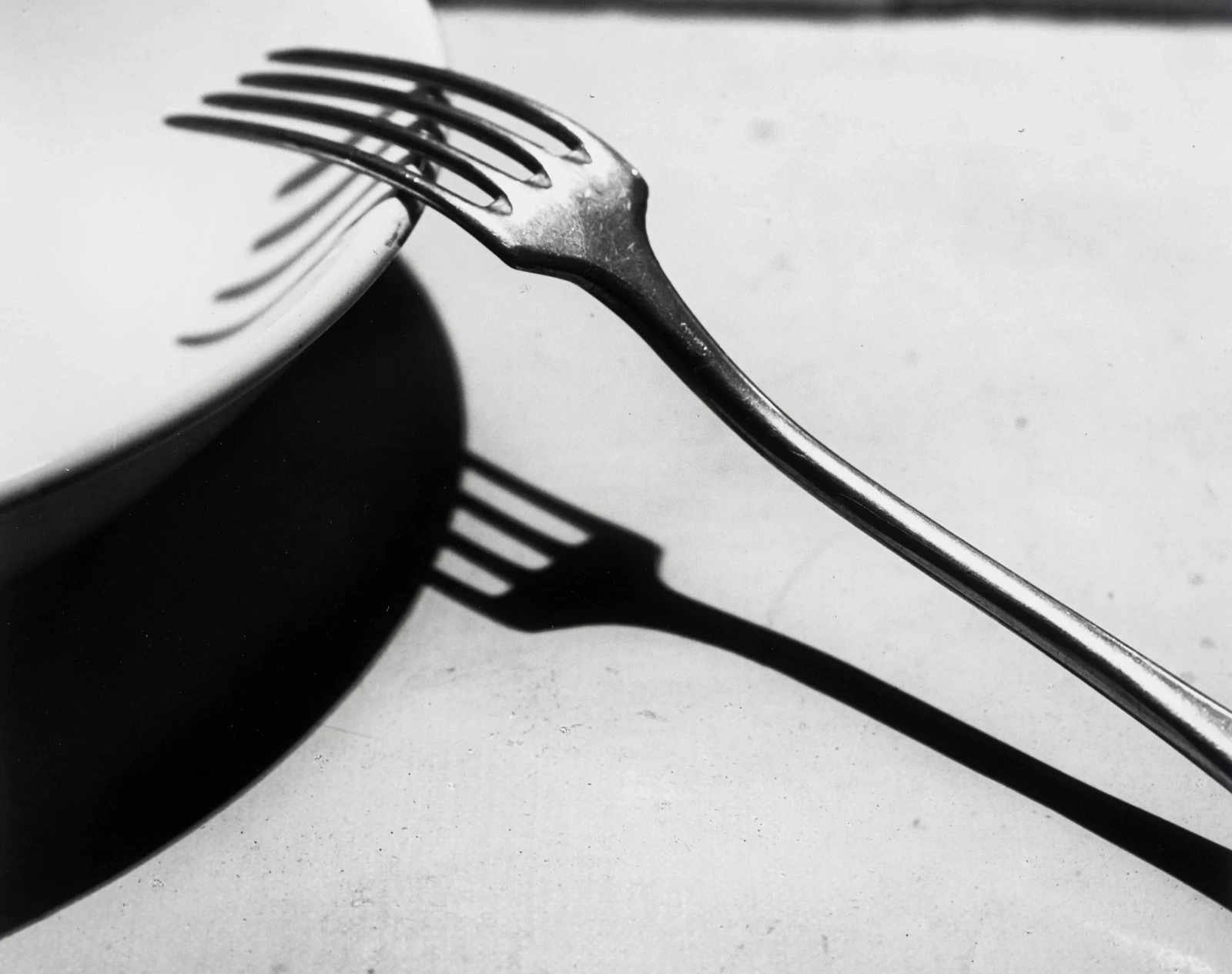 Black-and-white photograph of a fork leaning against the curved rim of a plate, with the fork’s dark shadow crossing the pale tabletop.
