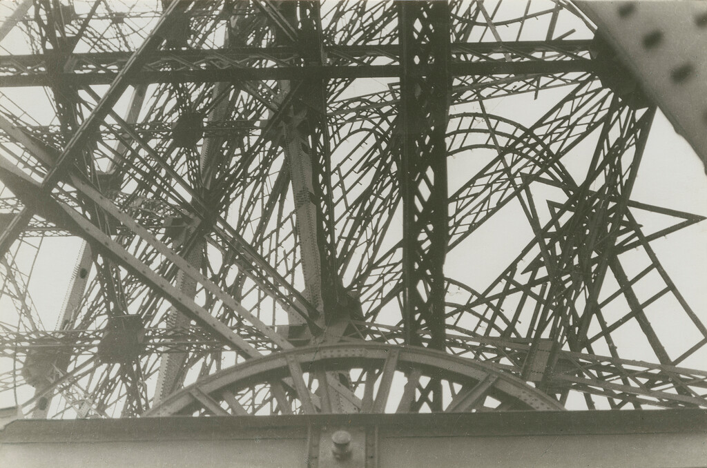 An upward view into the Eiffel Tower, with curved riveted beams crossing a dense lattice of diagonal iron braces.
