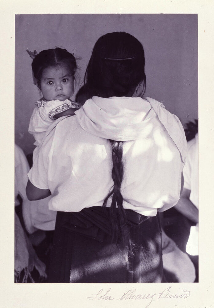 洛拉·阿尔瓦雷斯·布拉沃拍摄的黑白照片：一名女子背对镜头，长辫垂在浅色上衣上，怀里的孩子望向相机。