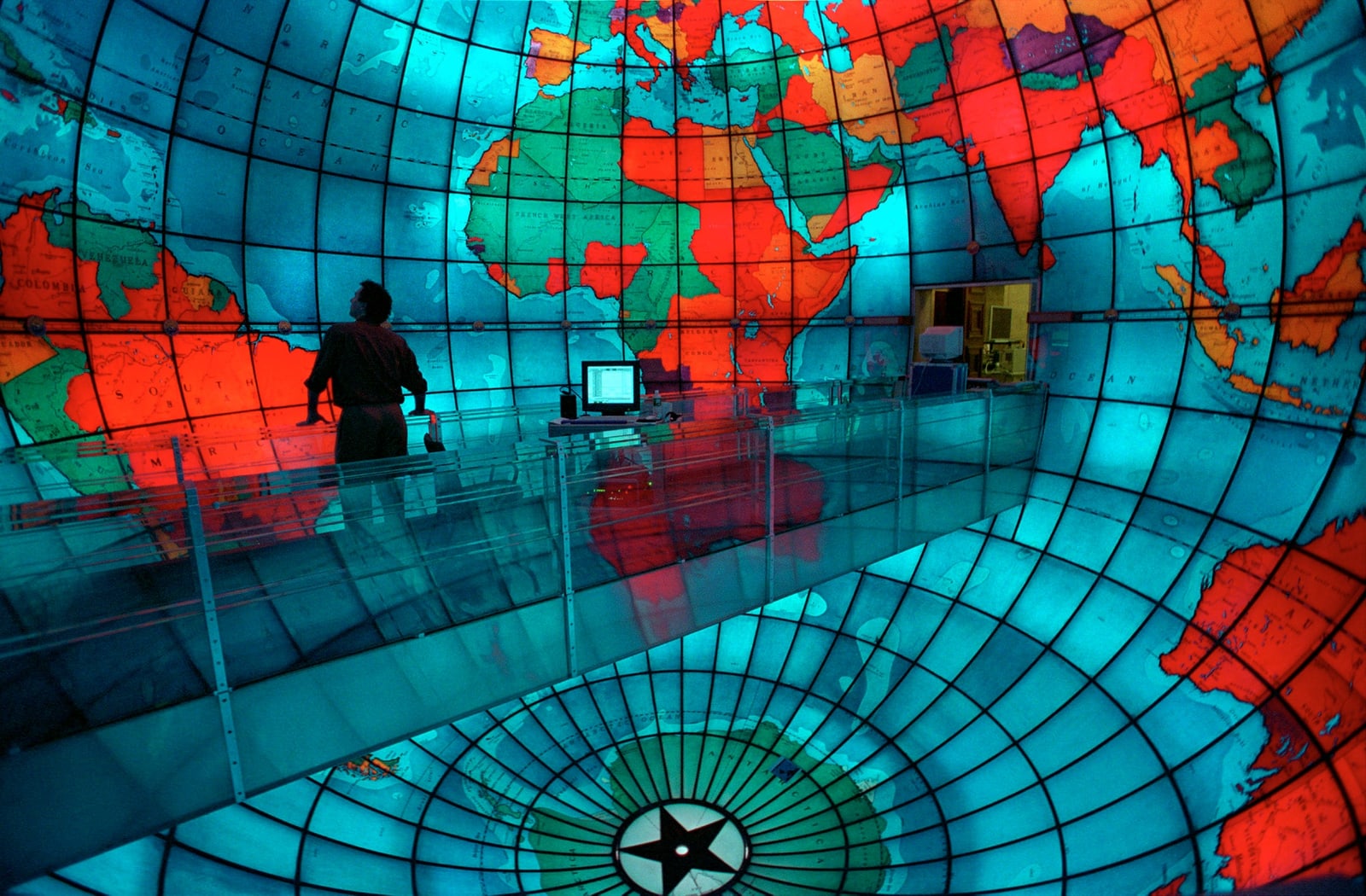 一位参观者站在波士顿 Mapparium 内的透明桥上，弧形玻璃板上发亮的蓝色海洋与色彩各异的国家从头顶到脚下环绕四周。