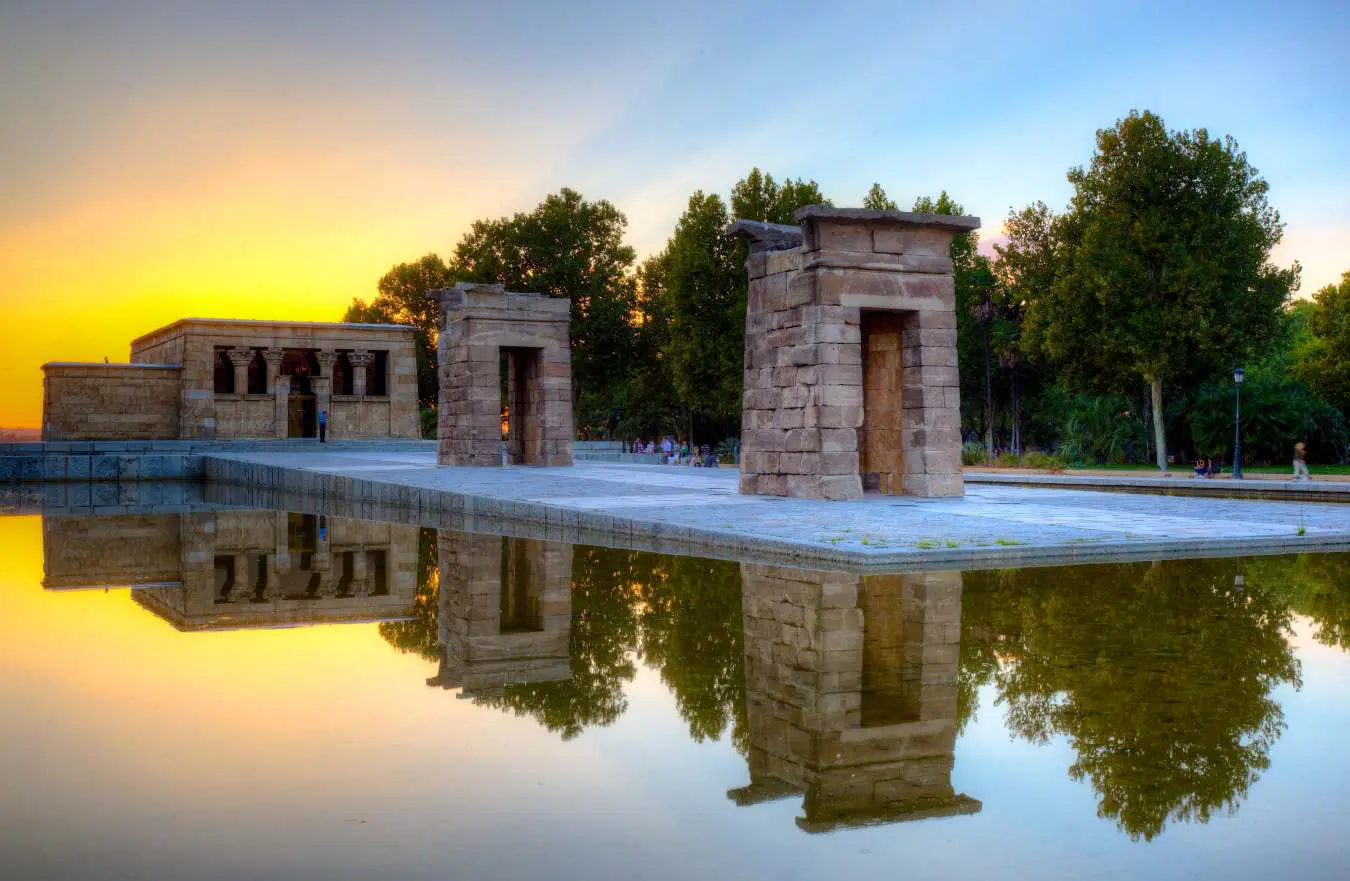 Two stone gateways and the Templo de Debod reflected in a pool beneath a yellow and blue evening sky in Madrid.