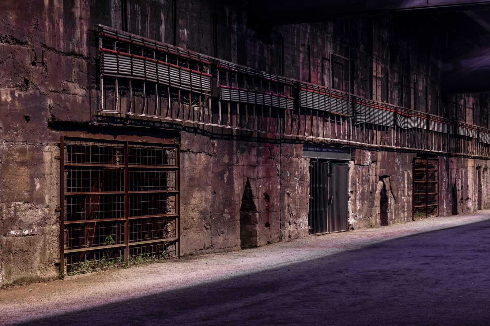 Pipes and barred openings run along a weathered industrial wall, lit in muted purple above the dark furnace street in Landschaftspark Duisburg-Nord.