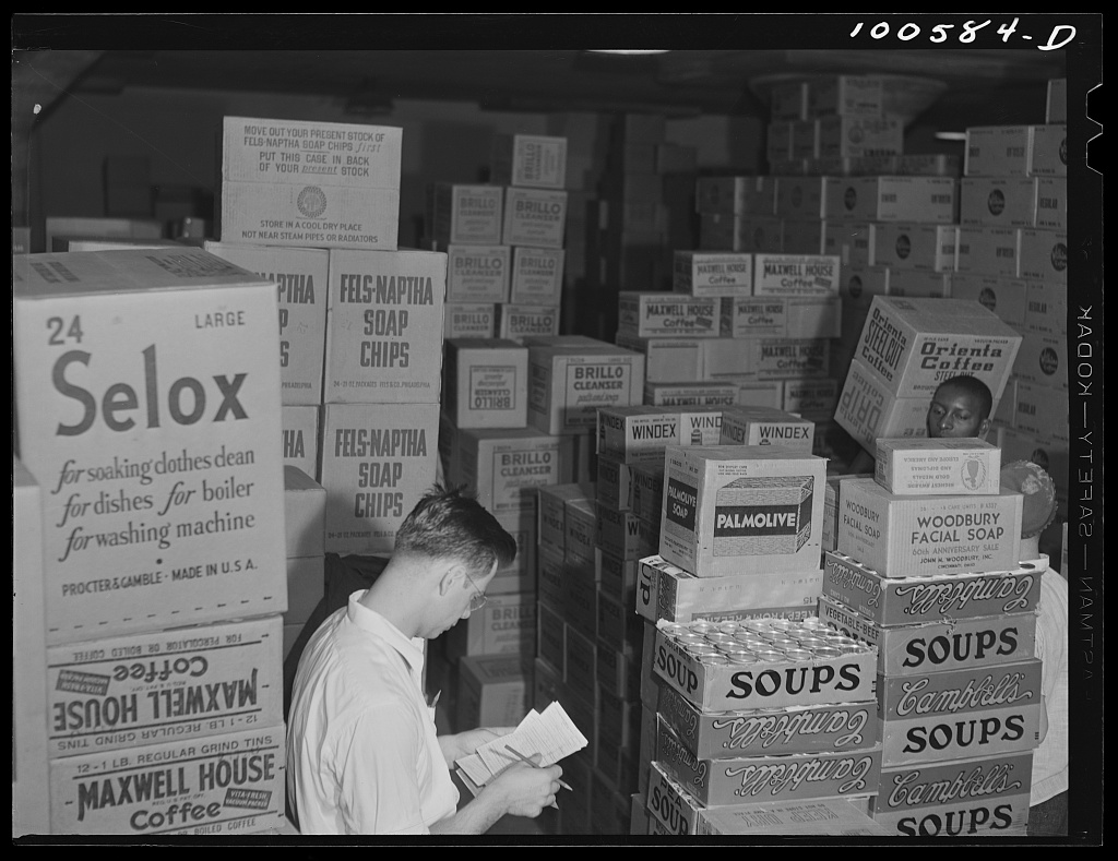 Workers checking merchandise among stacked cartons in the grocery department of a Washington, D.C., warehouse.