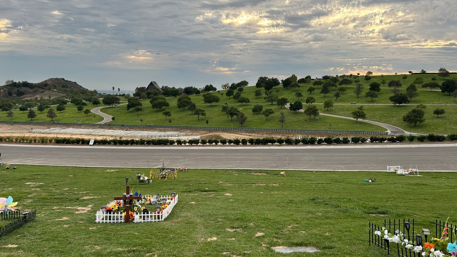 Green cemetery lawns dotted with flowers and trees, with a winding road and distant hills beneath a cloudy sky at Rose Hills Memorial Park.