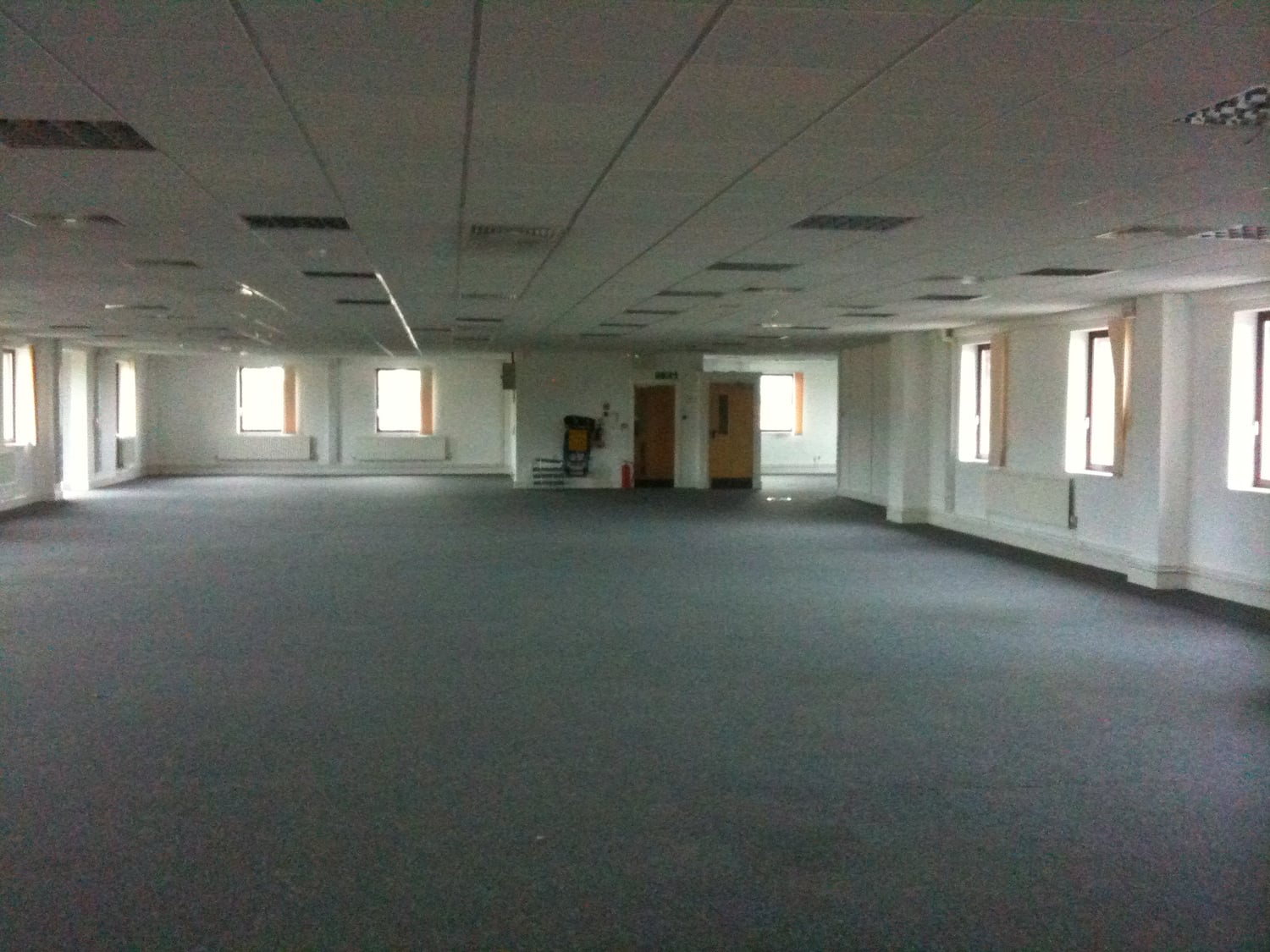 An empty office with grey carpet, white walls and daylight entering through the windows.