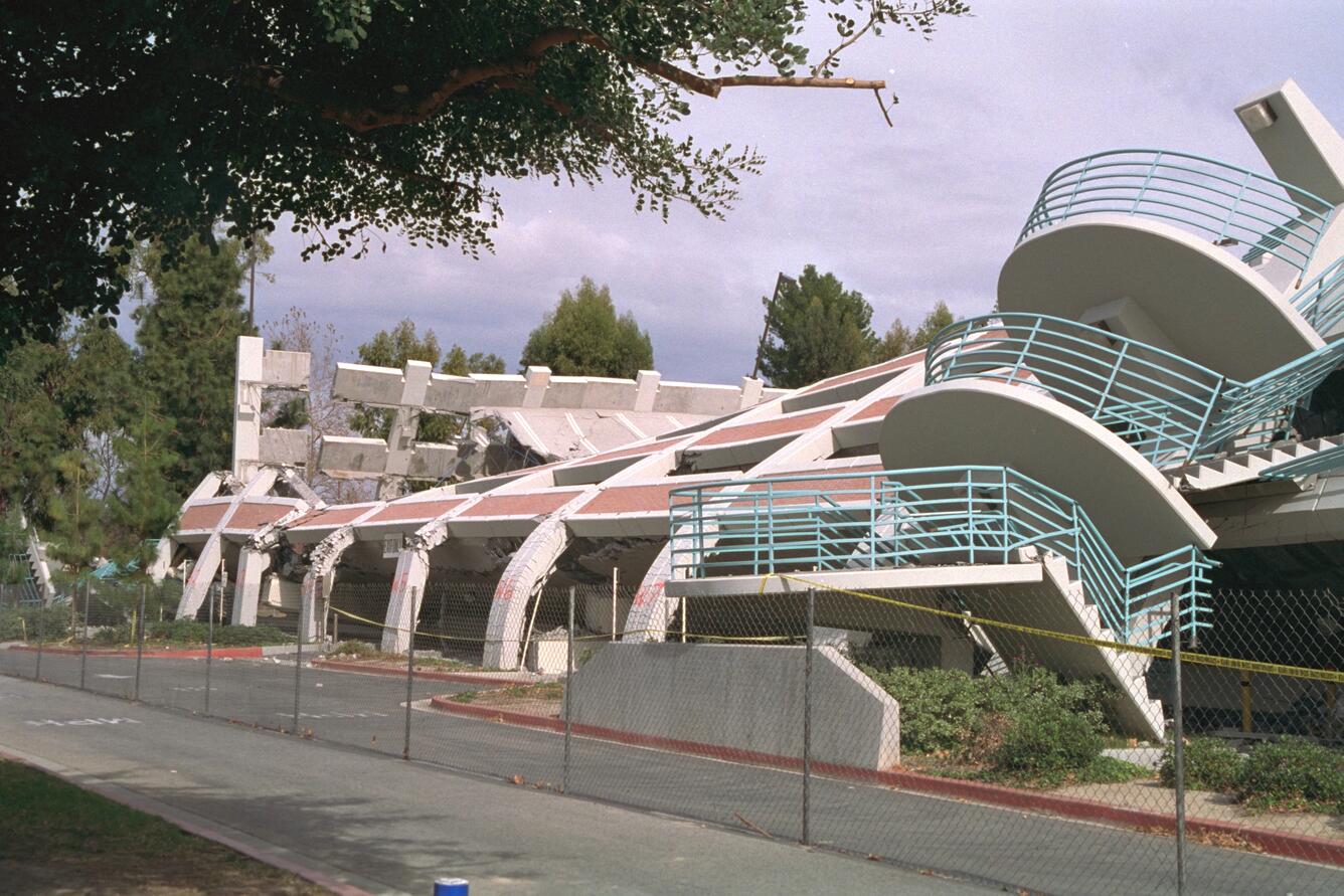 Collapsed concrete building behind a fence, with curved stair landings still visible, after the 1994 Northridge earthquake.