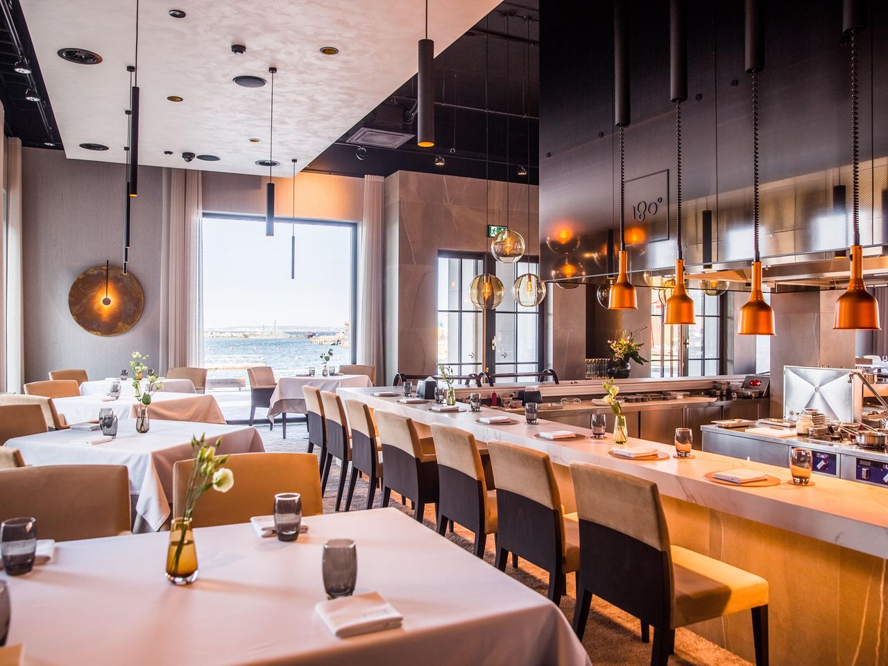Cream chairs and white-clothed tables surround the open kitchen at 180°, with copper lamps above the counter and Tallinn's harbour visible through a large window.