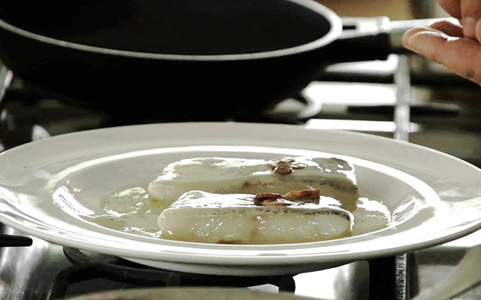 Two portions of skin-on cod coated in pale pil-pil sauce on a white plate beside a cooking pan.