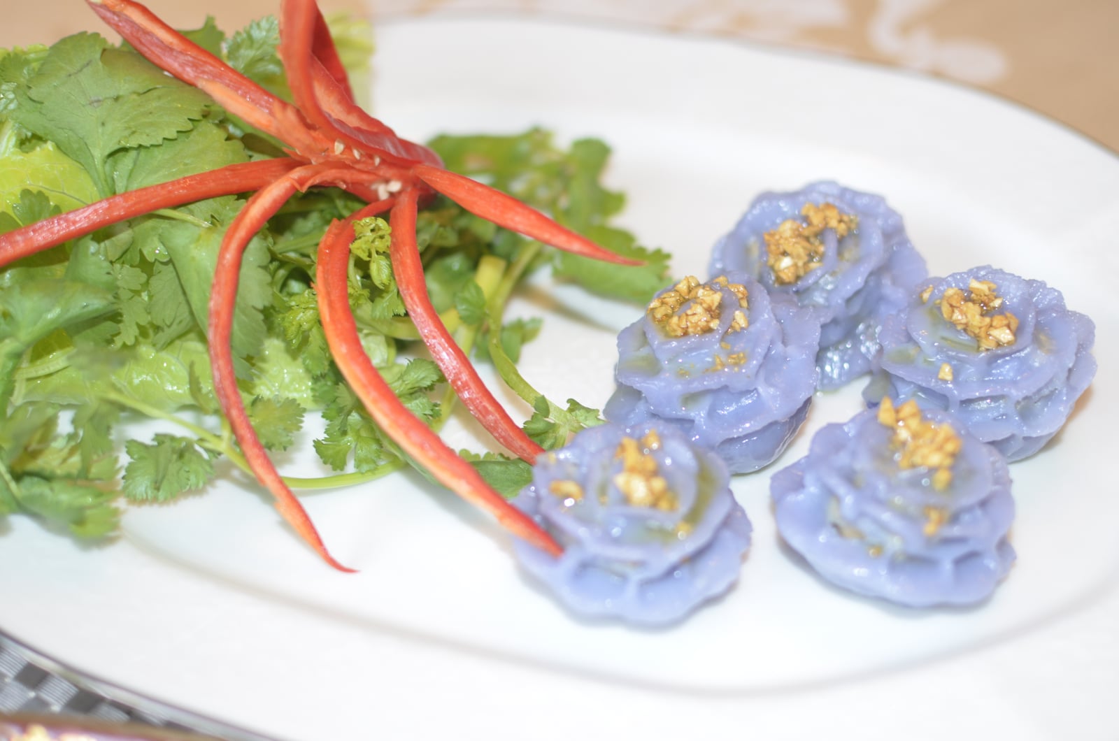 Five purple chor muang dumplings topped with fried garlic, beside coriander leaves and red chile strips on a white plate.