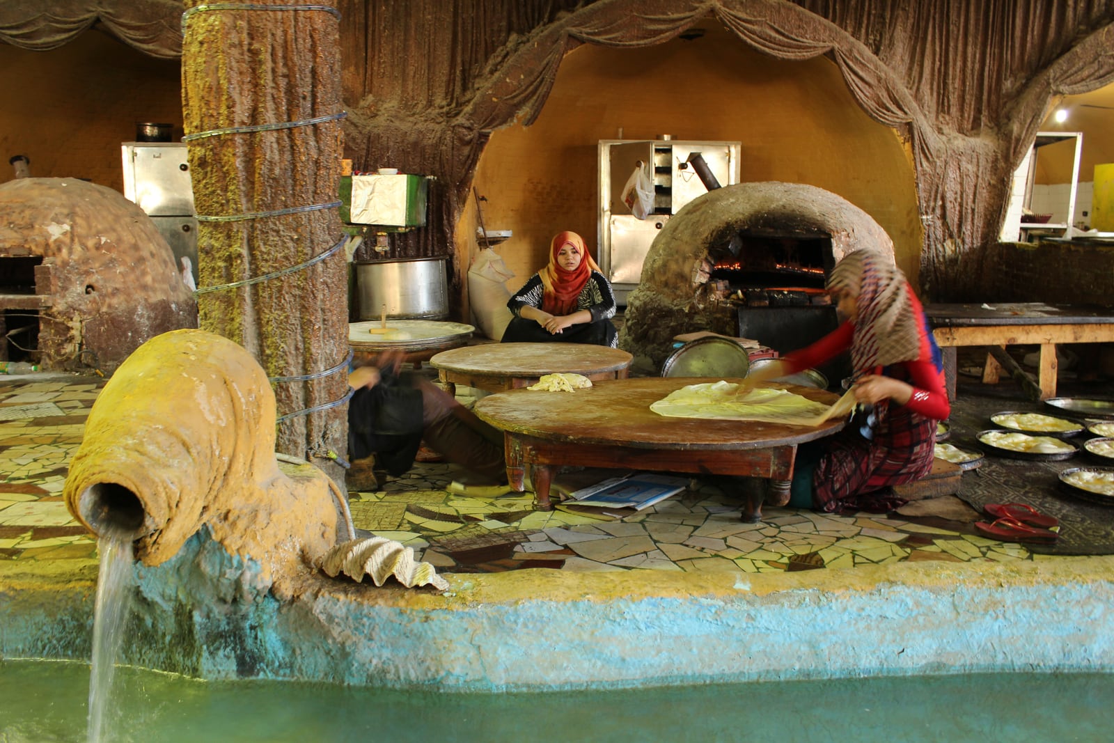 A woman stretches feteer dough over a round wooden table beside domed ovens and trays of unbaked pastry.