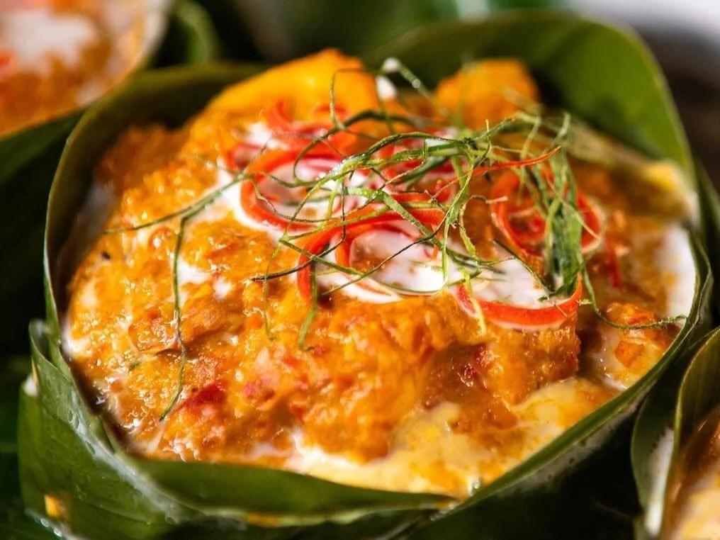 A close view of fish amok in a folded banana-leaf cup, topped with coconut cream, red pepper curls and finely sliced lime leaves.
