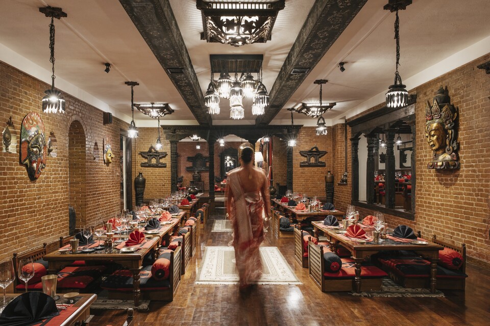 Krishnarpan 的砖墙餐室里，雕花木梁下，一名工作人员从摆齐餐具的矮桌与红色坐垫之间走过。