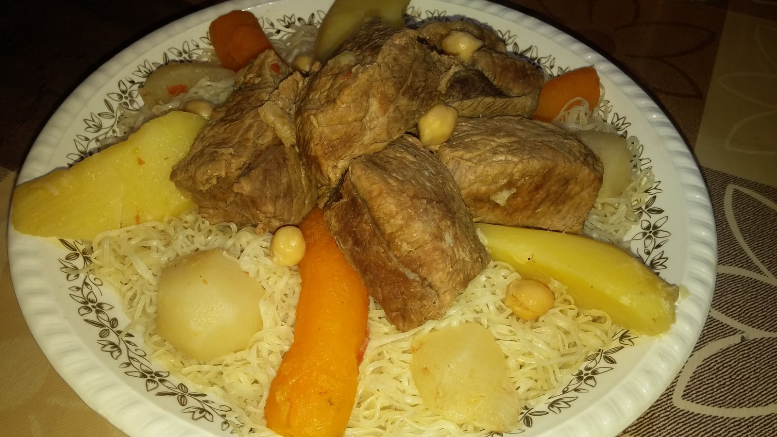 Large pieces of meat, chickpeas and vegetables on fine rechta noodles in a patterned platter.