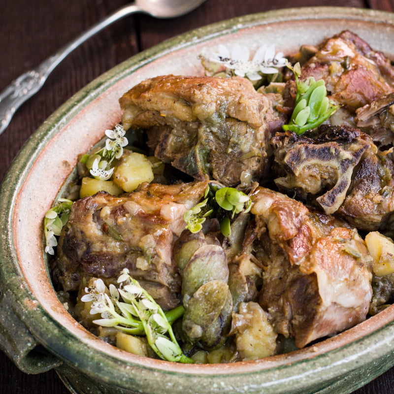 Browned lamb pieces, potatoes, and green waterblommetjies in a ceramic casserole, garnished with white blossoms.