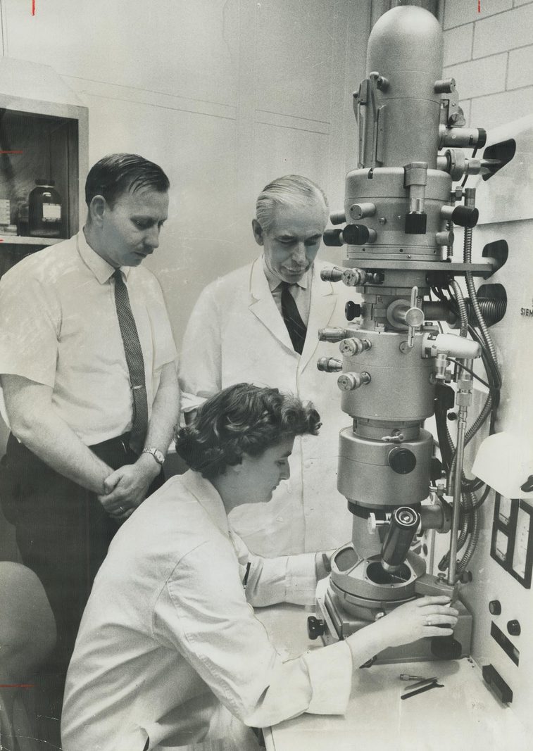 June Almeida in a white laboratory coat operating an electron microscope, with two men observing beside her.