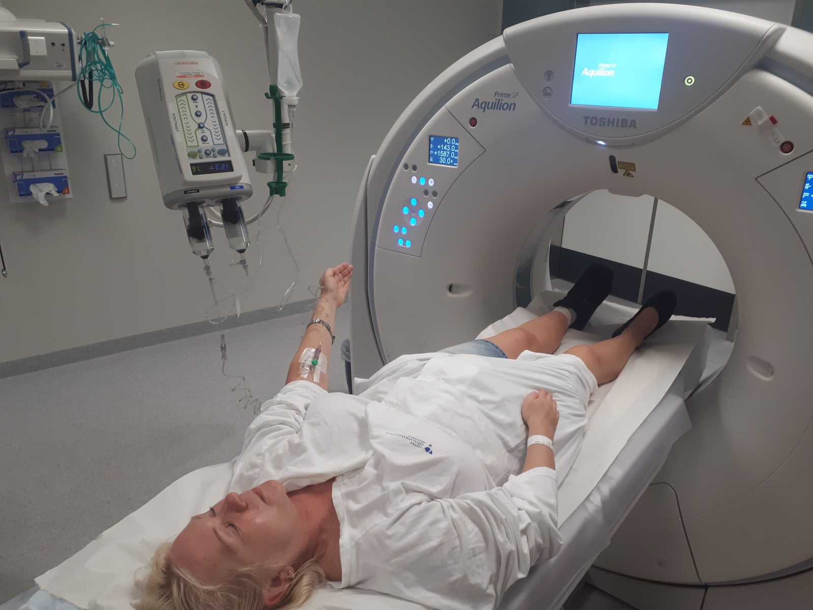 A patient lies on a CT scanner table with an intravenous line connected to the contrast delivery equipment at the upper left.