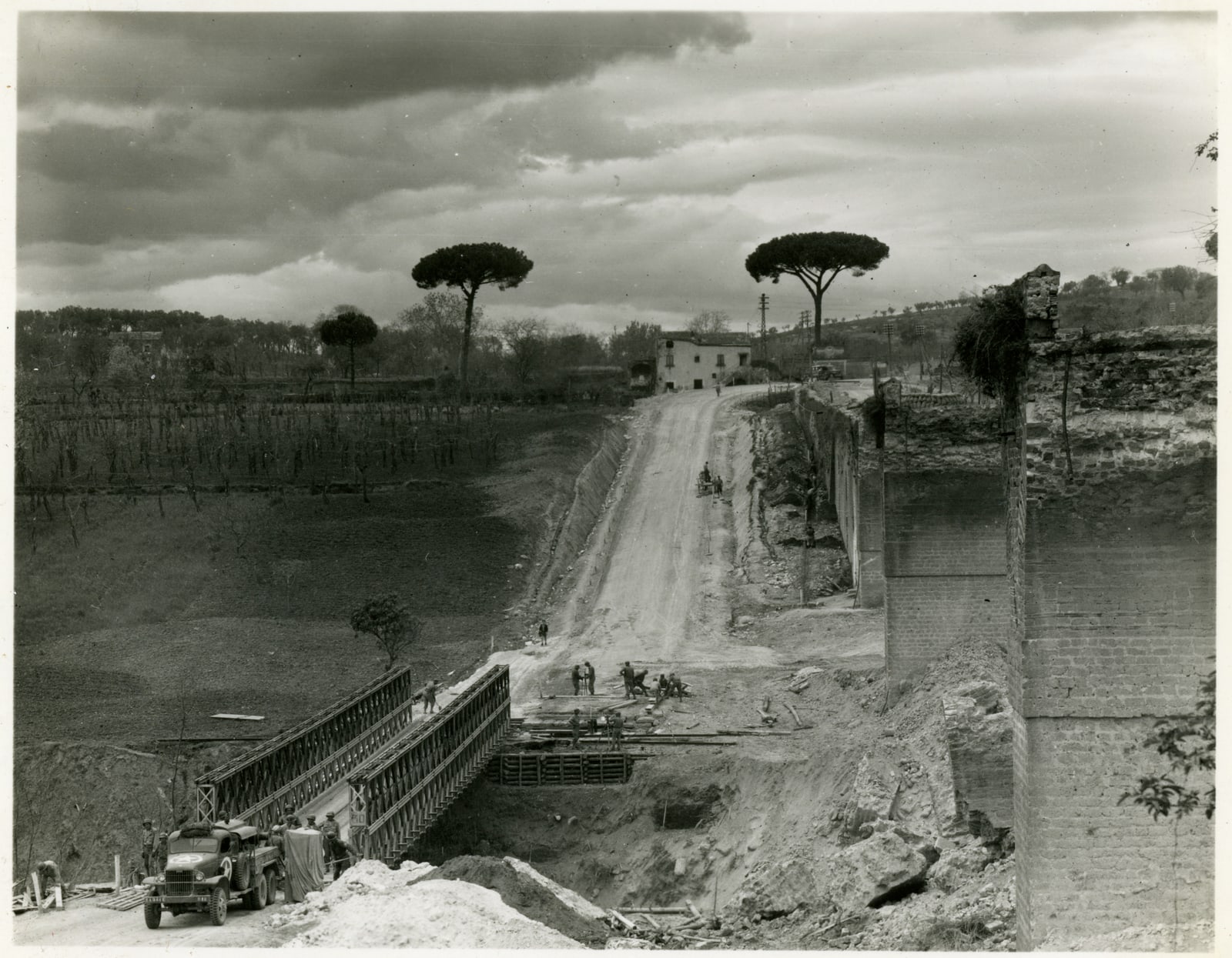 American engineers beside a steel Bailey bridge, with a truck in the foreground, ruined masonry to the right, and a road climbing into the Italian countryside.