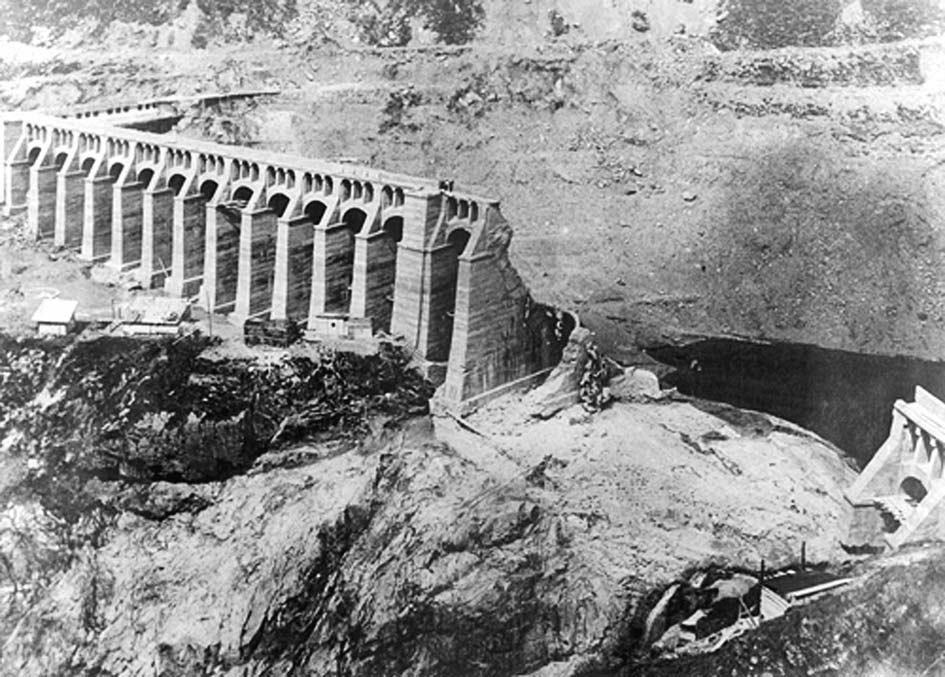 Archival photograph of Gleno dam after the collapse, with a row of surviving buttresses ending beside the broad central breach and exposed masonry below.