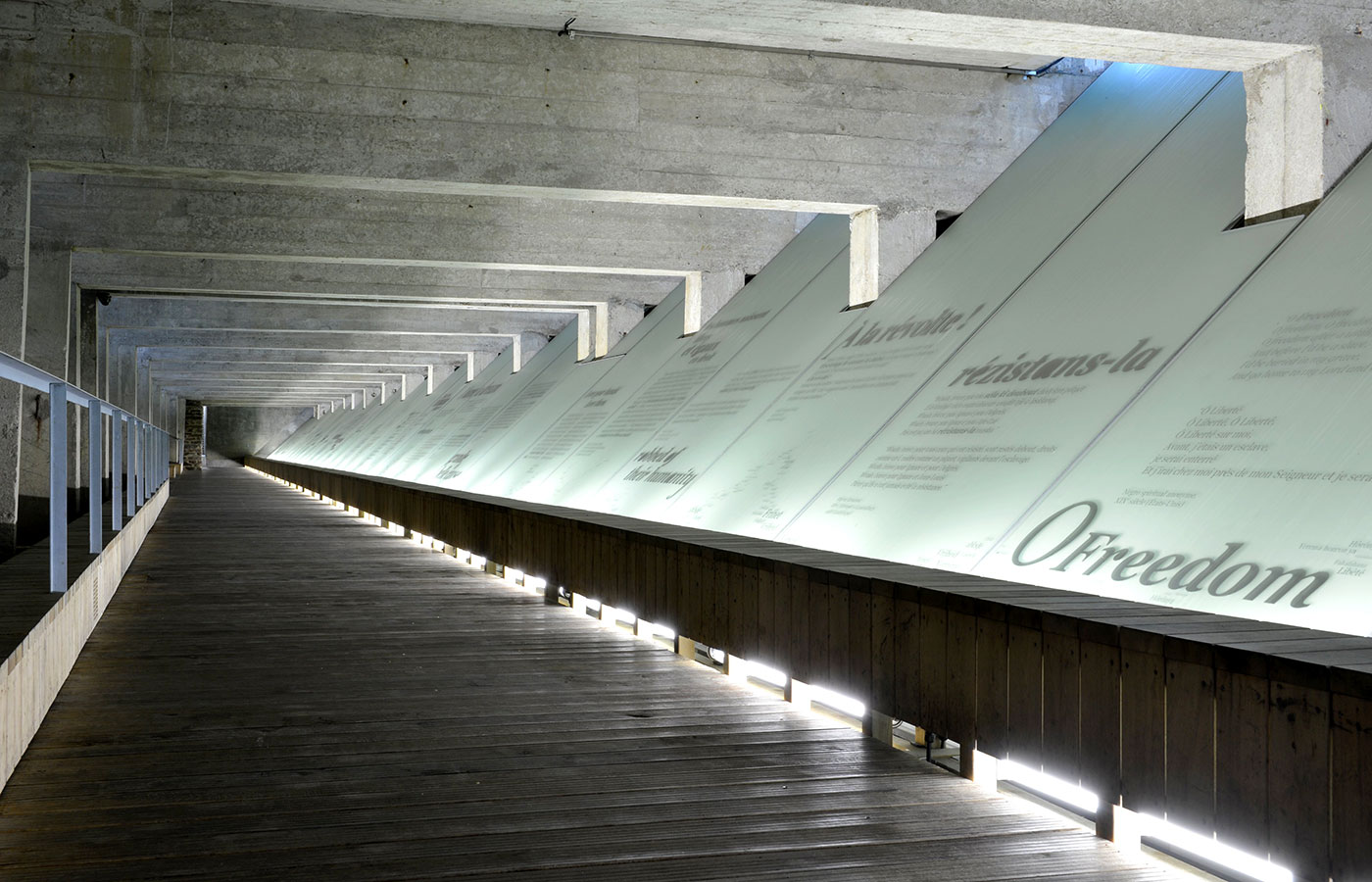 A long timber walkway beneath concrete beams, with illuminated sloping glass panels bearing texts about freedom at the Nantes memorial.