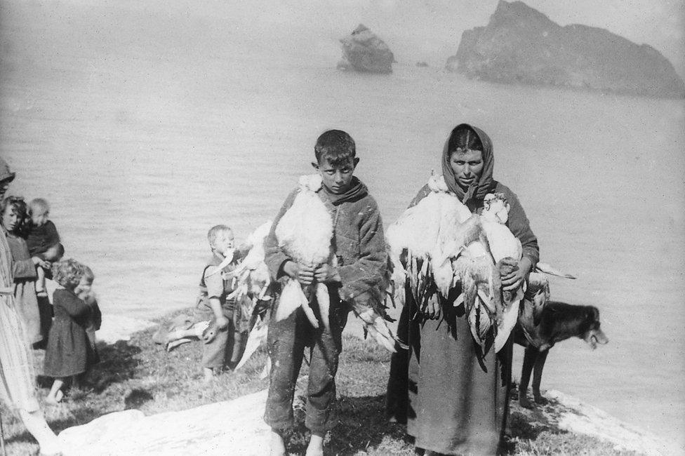 Black-and-white photograph of a woman and a boy holding strings of seabirds beside a rocky shore, with children and a dog nearby.