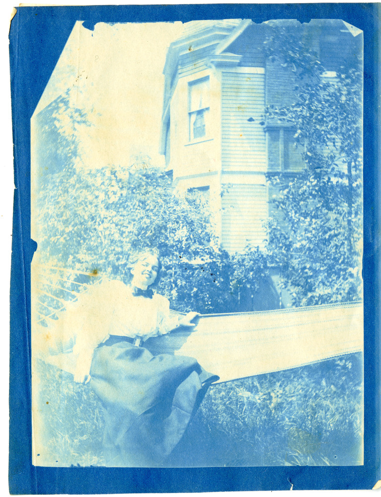 Blue cyanotype of Adelaide Crapsey smiling in a hammock outside a wooden house, wearing a white blouse and long dark skirt.